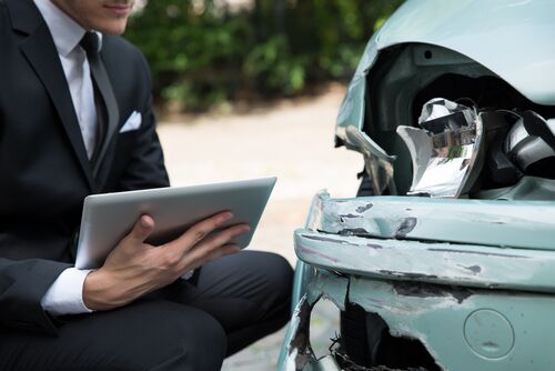 Man in a suit holding a tablet while inspecting damage to the front of a dented car.
